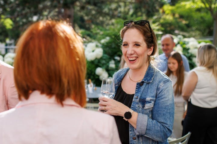 Frau in Jeansjacke hält ein Glas und unterhält sich lächelnd im Freien.