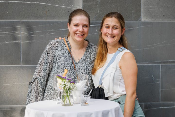 Zwei lächelnde Frauen an einem Tisch mit Blumendeko und Gläsern bei einer geselligen Veranstaltung.