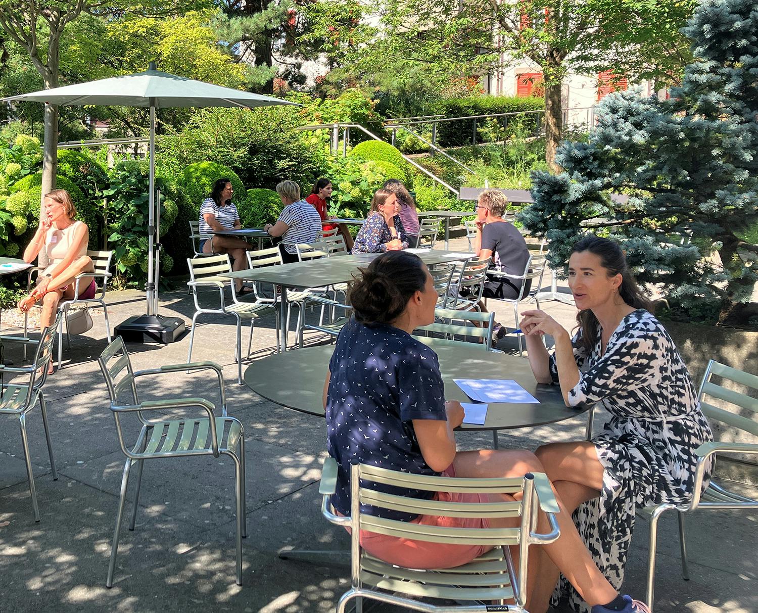 Personen sitzen im Freien in einem Café mit Sonnenschirmen umgeben von Grünanlagen.