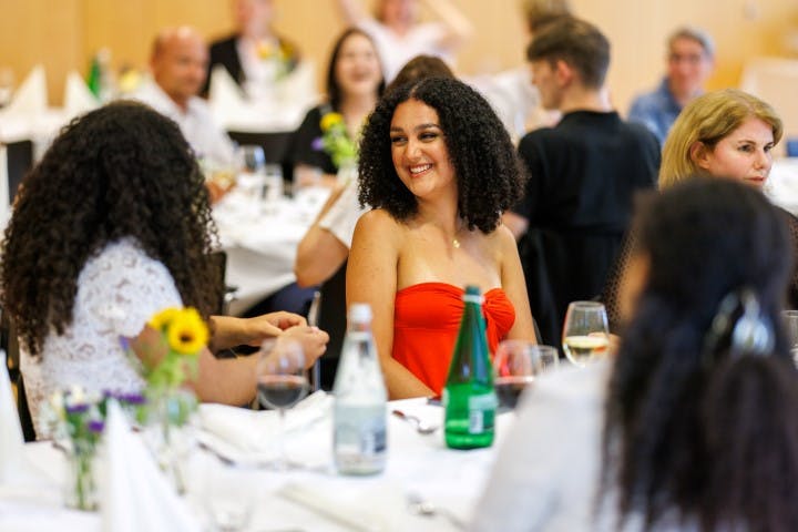 Frau in rotem Oberteil lächelt bei einer geselligen Veranstaltung mit Gästen am Tisch.