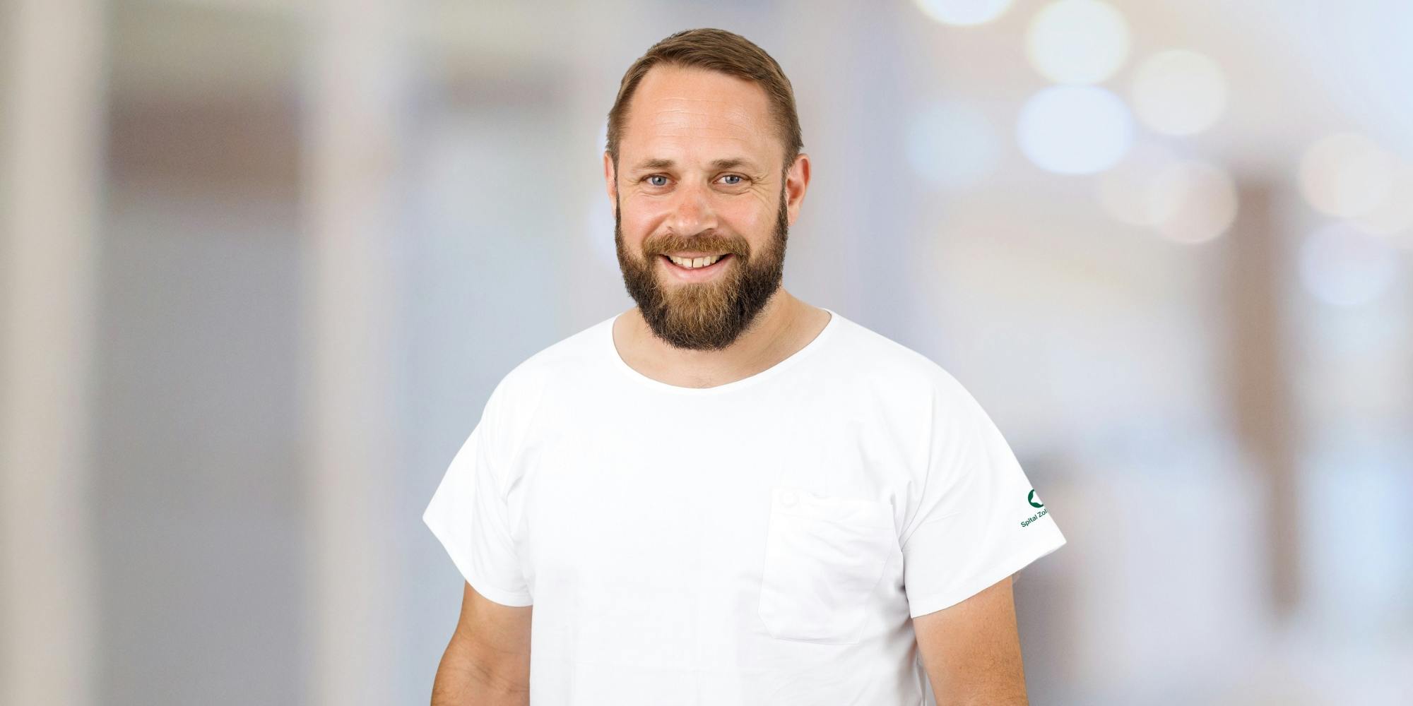 Lächelnder Mann mit Bart in weißem T-Shirt vor unscharfem Hintergrund.