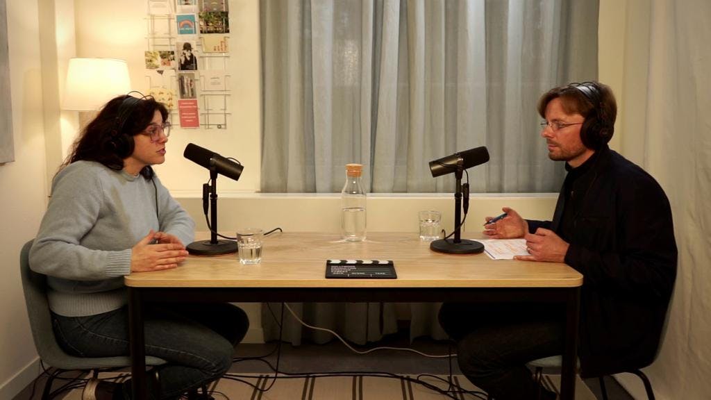 Zwei Personen sitzen an einem Tisch im Gespräch mit Mikrofonen für eine Podcast-Aufnahme.