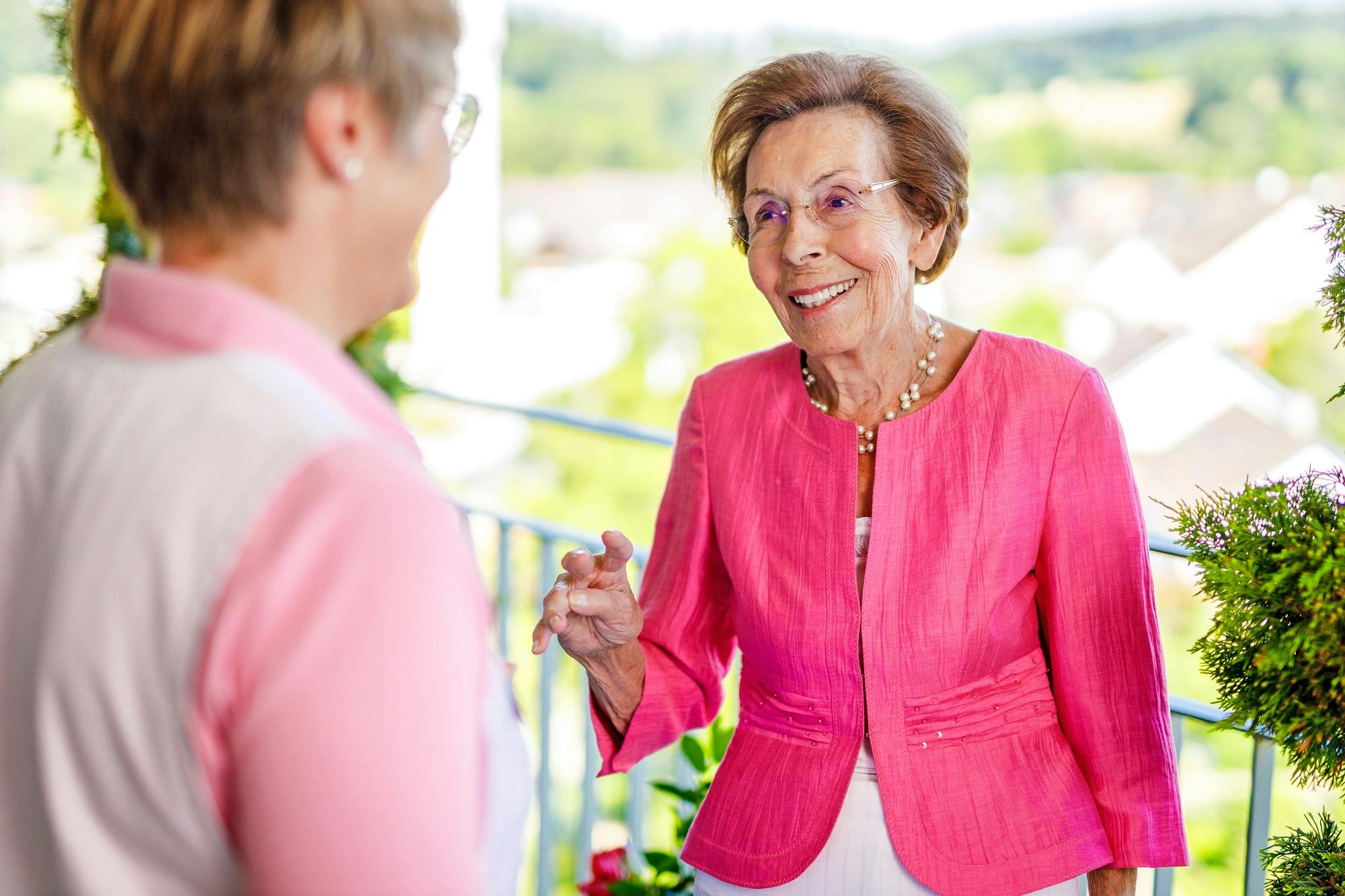 Ältere Dame in rosa Jacke unterhält sich lächelnd auf einem Balkon.