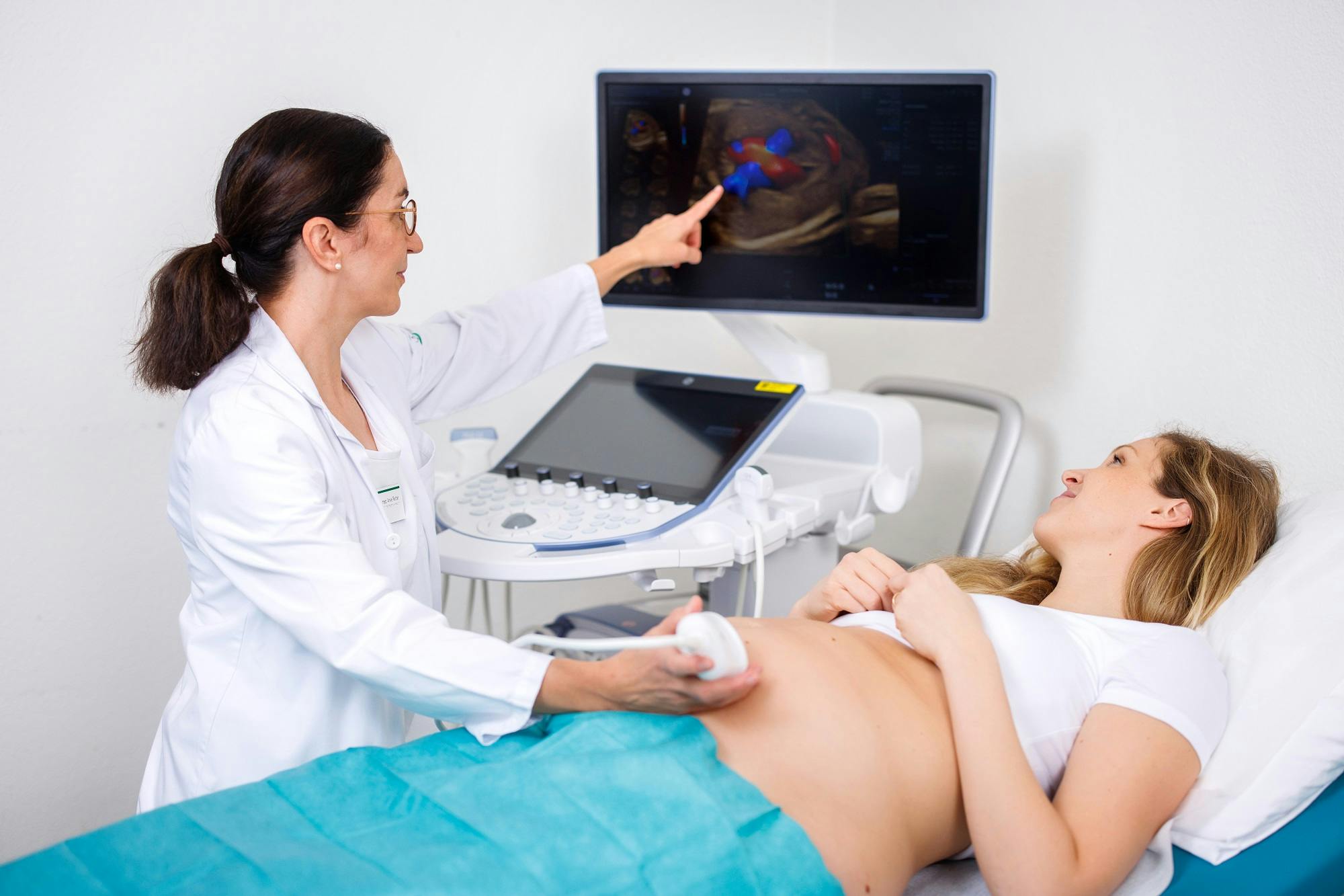 Ärztin führt Ultraschalluntersuchung bei Patientin durch und deutet auf Ultraschallbild am Monitor.