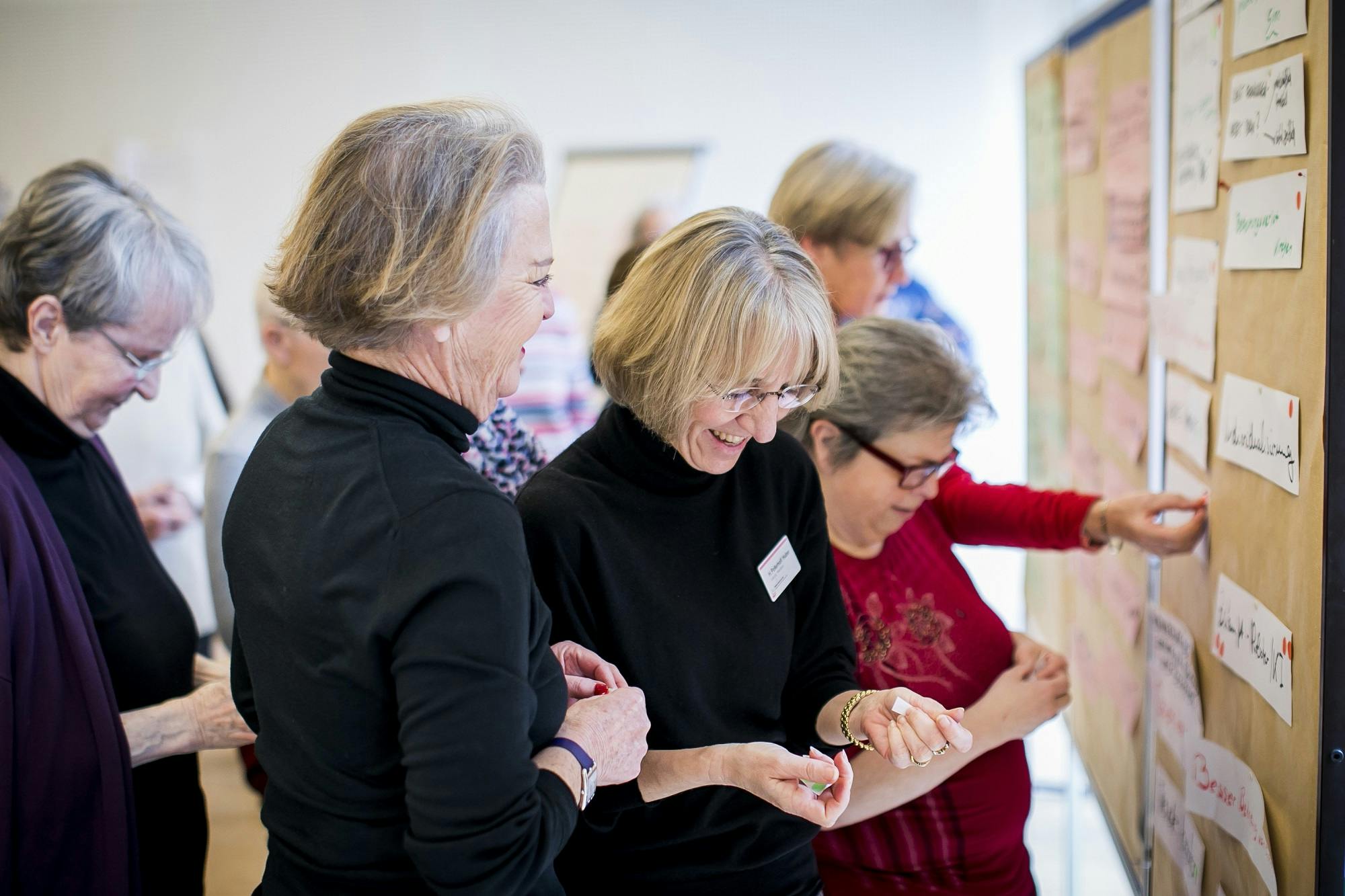 Gruppe älterer Frauen, die gemeinsam lachen und auf einer Veranstaltung scheinbar aktiv eine Wandpräsentation betrachten.
