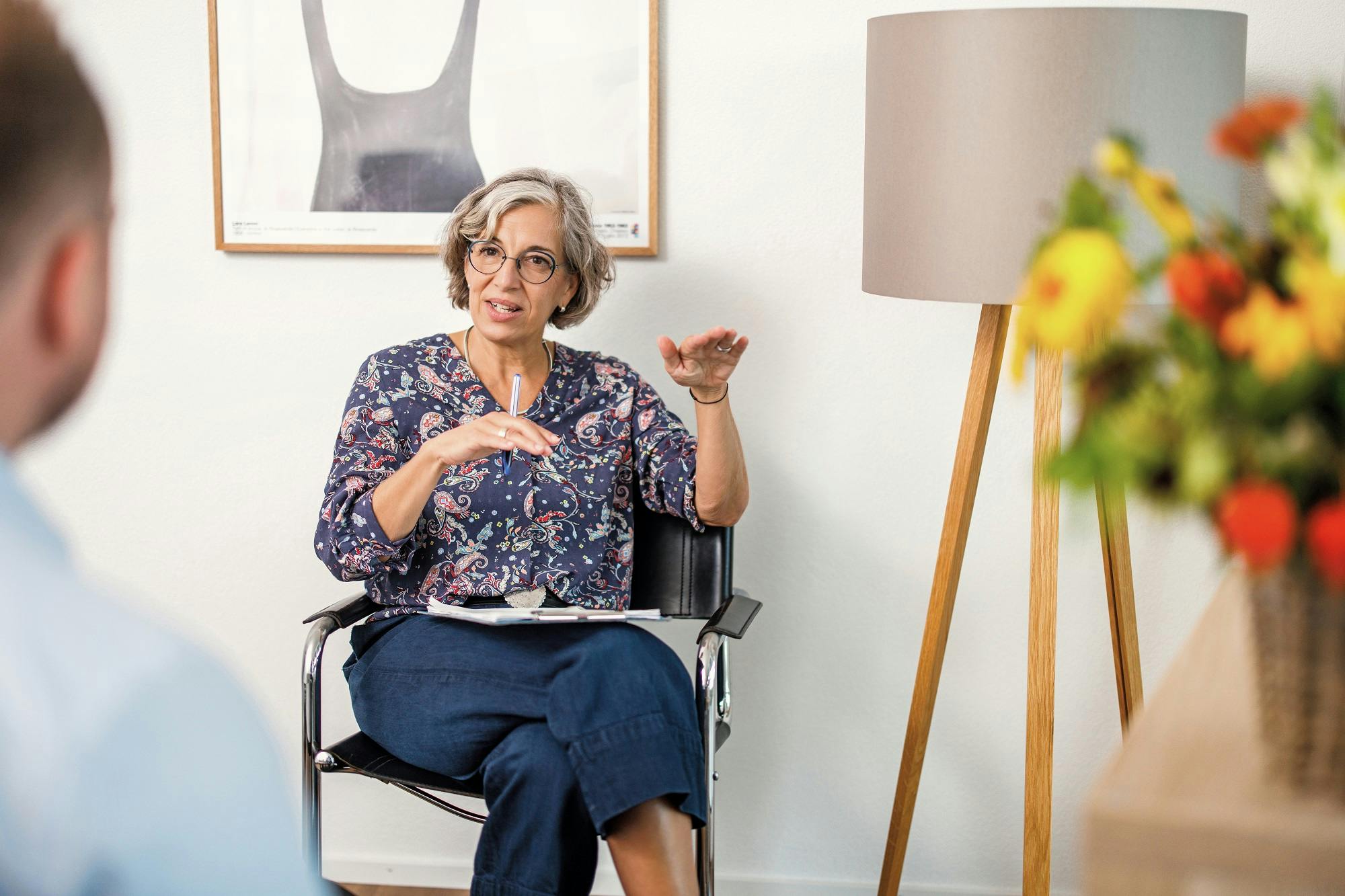 Frau im Rollstuhl mit Brille gestikuliert beim Sprechen in einem hellen Raum.