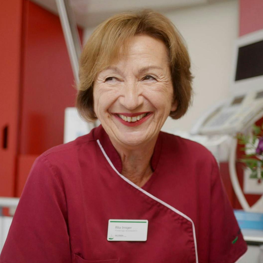 Lächelnde ältere Krankenschwester in roter Uniform mit Namensschild in einer Klinik.