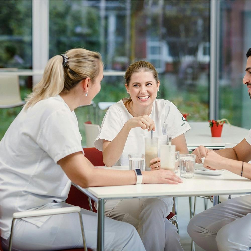 Drei lächelnde Personen in weißer Berufskleidung, die an einem Tisch sitzend, Getränke genießen und sich unterhalten.