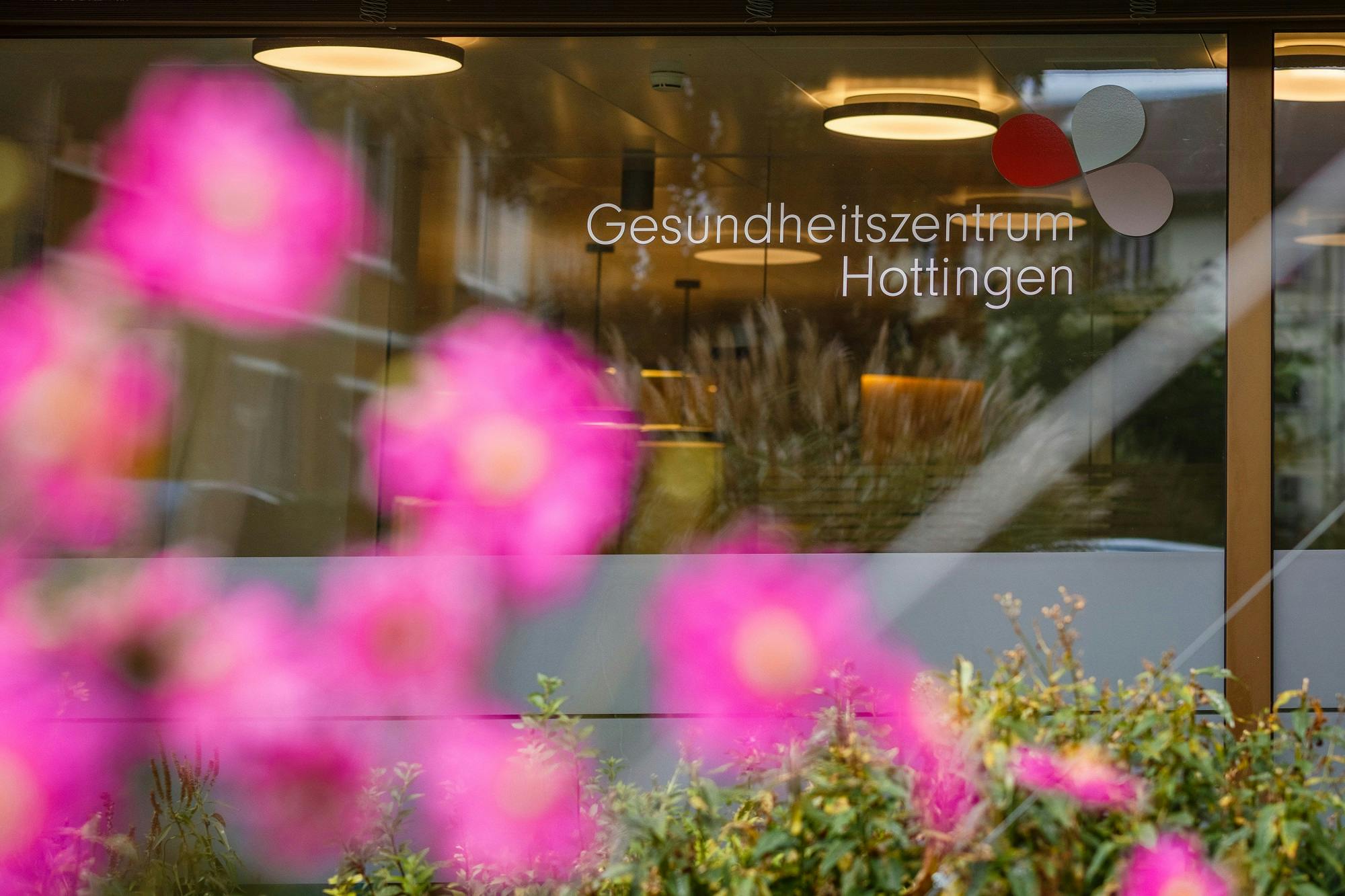 Schaufenster des Gesundheitszentrums Hottingen mit unscharfen rosa Blumen im Vordergrund.