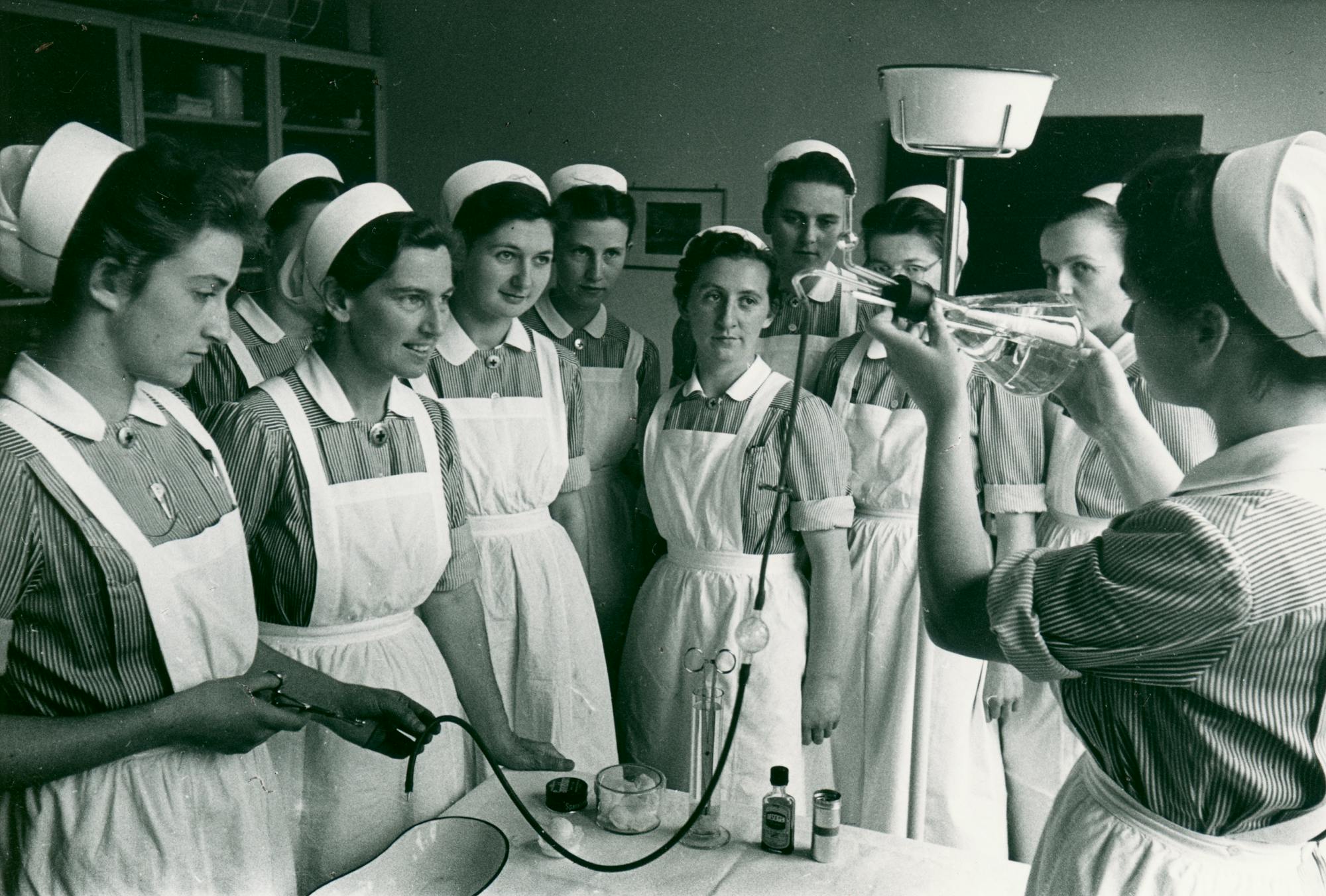 Gruppe von Krankenschwestern in traditioneller Uniform beobachtet medizinische Demonstration, Schwarz-Weiß-Fotografie.