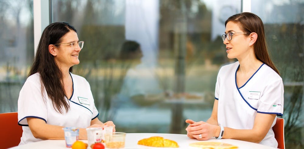 Zwei Personen in medizinischer Berufskleidung im Gespräch an einem Tisch mit Lebensmitteln.