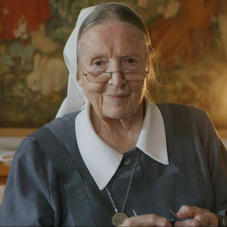 Ältere Nonne mit Brille und traditioneller Tracht lächelt vor einem gemalten Hintergrund.