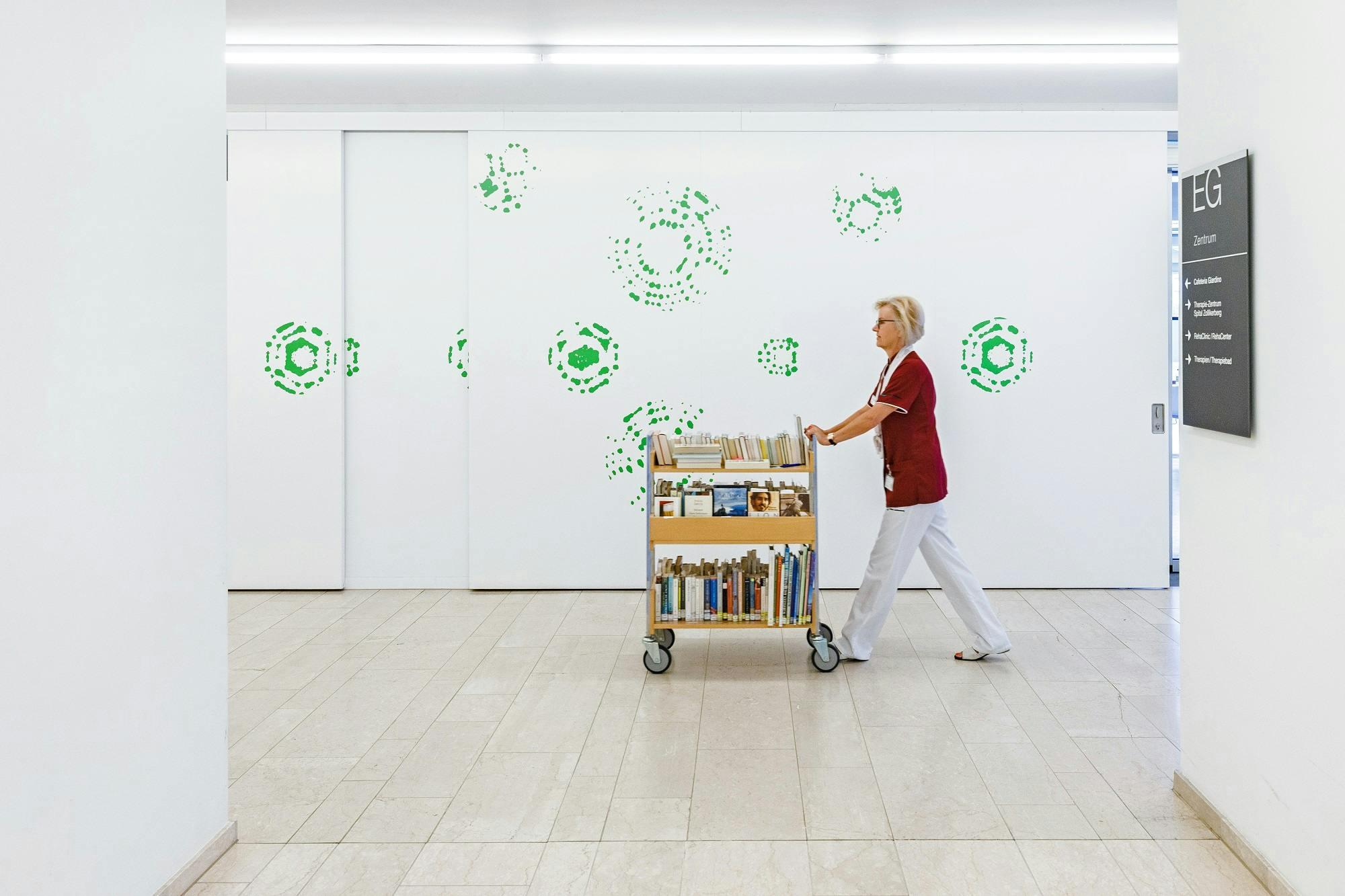 Frau schiebt Bücherwagen in einer Bibliothek mit grünen Wanddekorationen.