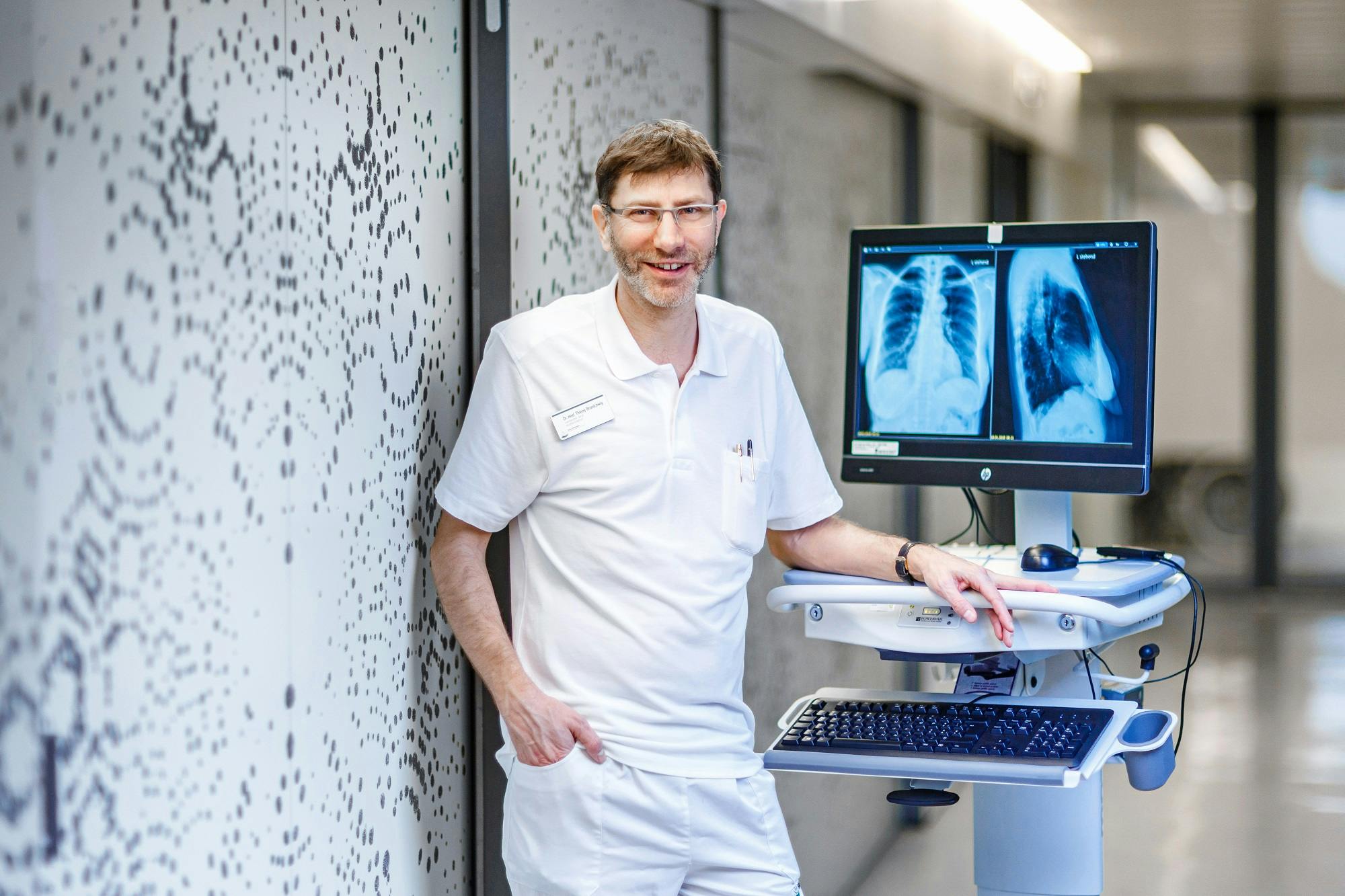 Lächelnder Arzt im weißen Kittel an einem Computer mit Röntgenbildern auf dem Monitor.
