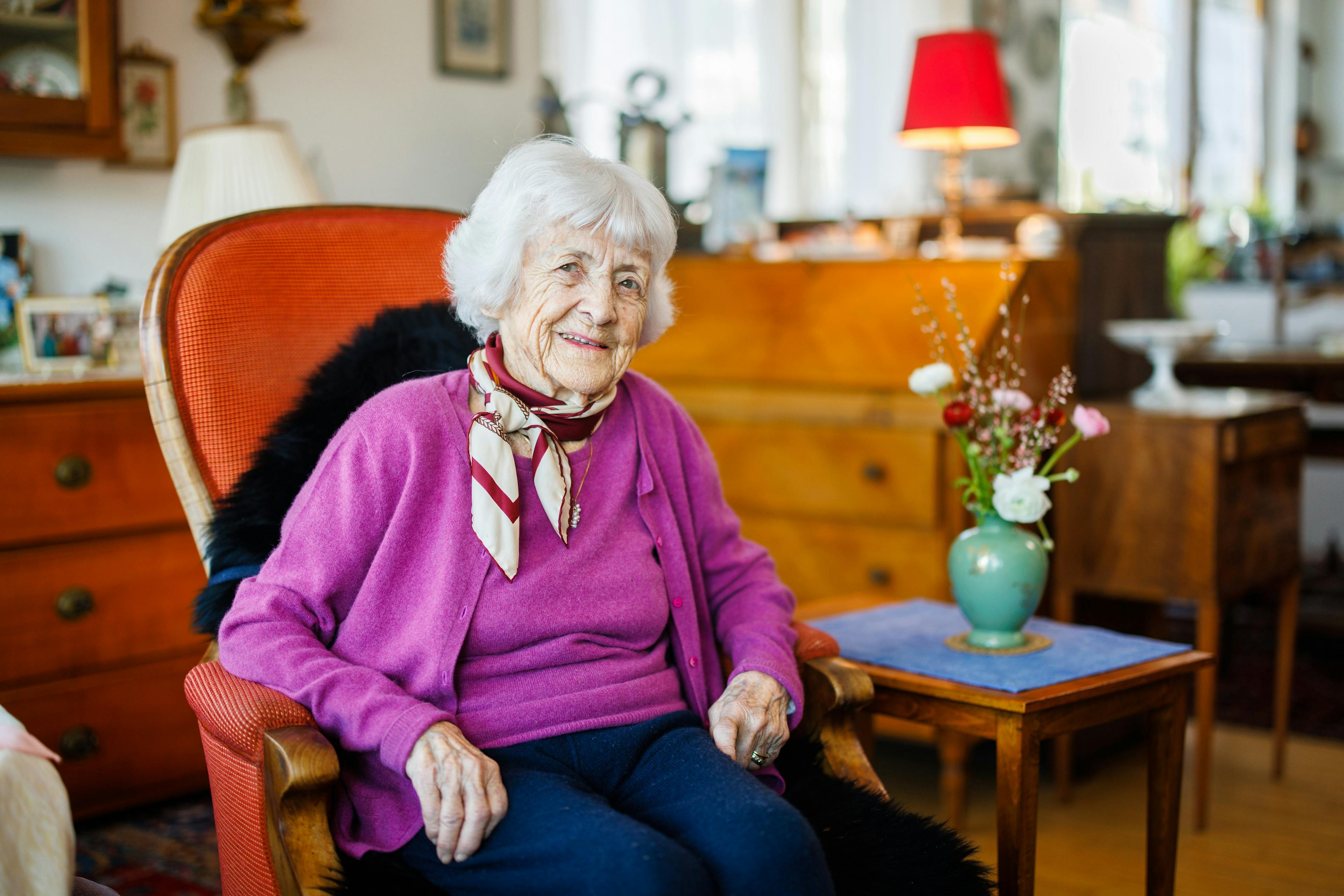 Eine ältere Dame sitzt lächelnd auf einem Stuhl in einem gemütlichen Wohnzimmer.