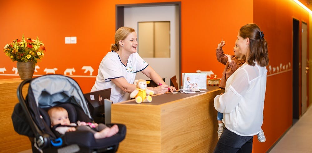 Empfangsdame im medizinischen Kittel begrüßt lächelnd eine Person an einem orangefarbenen Rezeptionstresen.